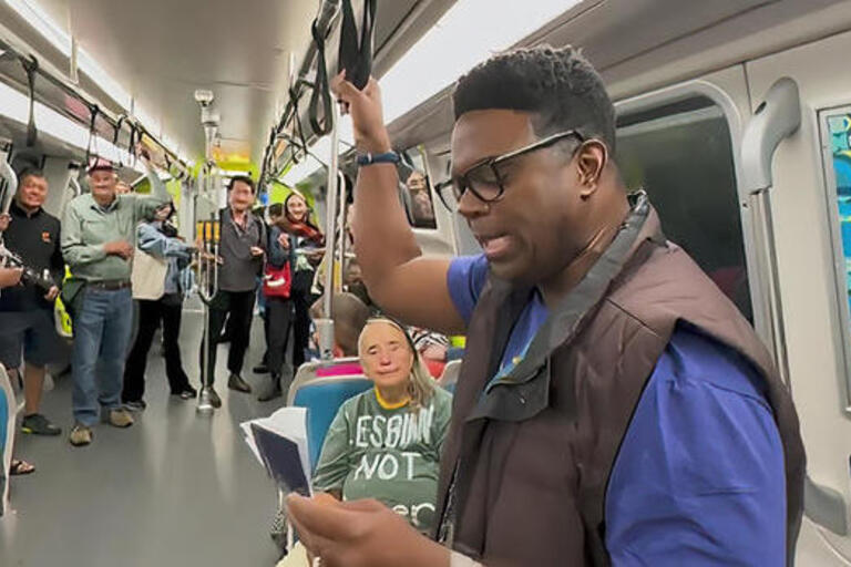 a person standing in a BART car reads aloud from an open book while other passengers smile