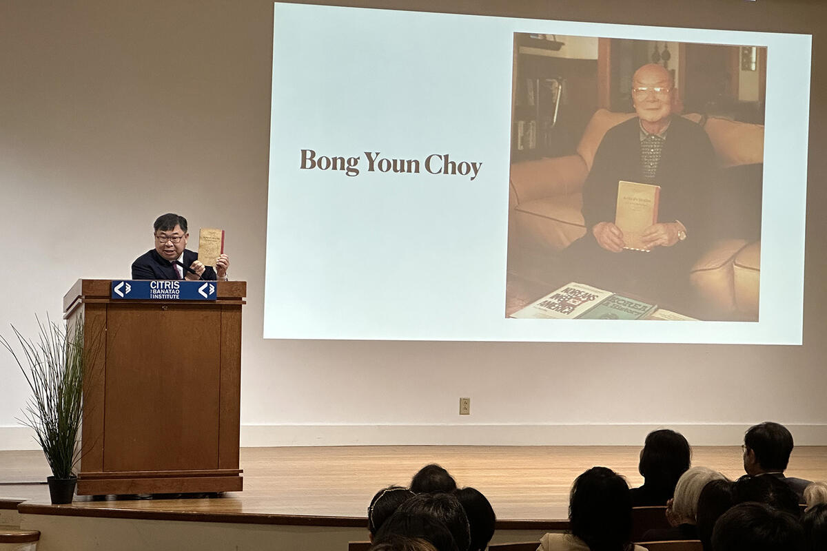 A man at a lectern holds a copy of Korean Reader-A Textbook for Beginners in front of a projection of Bong Youn Choy holding the same book