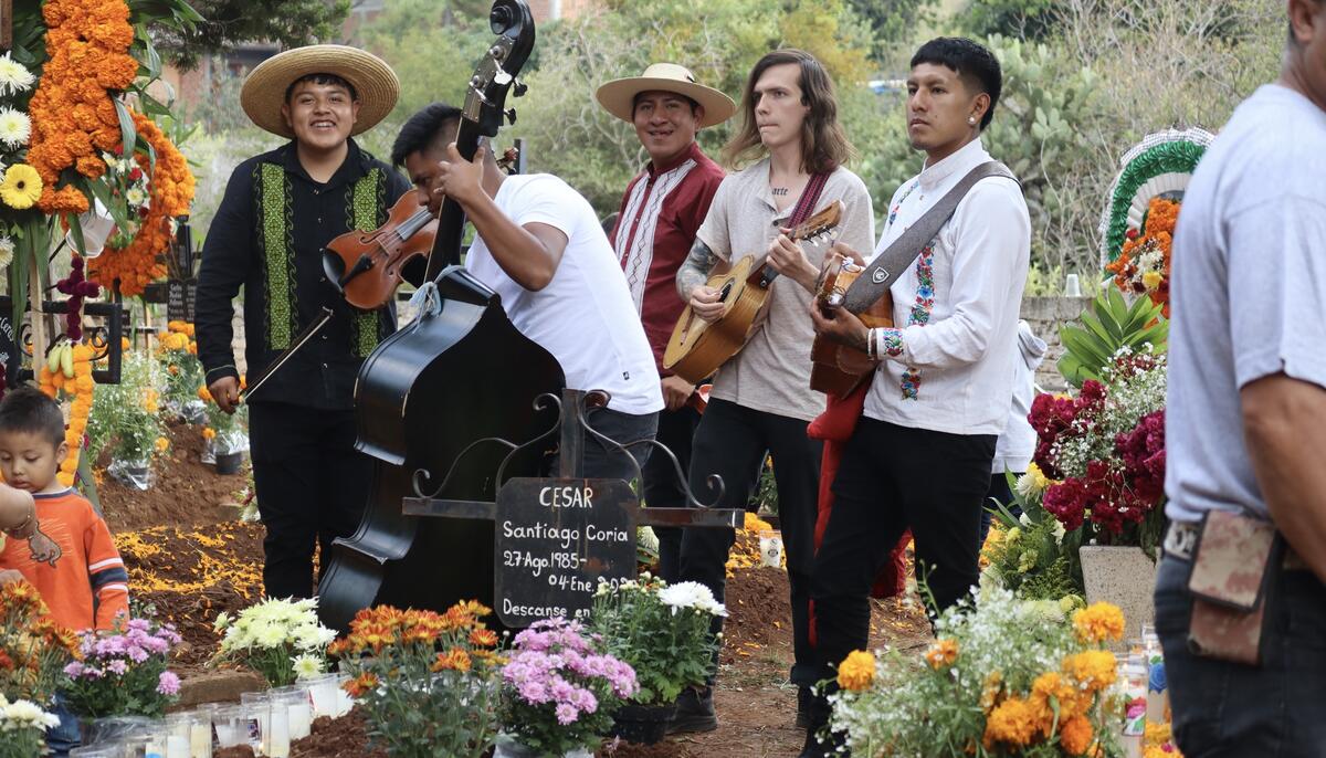 Musicians performing at a decorated grave in a cemetery with vibrant floral arrangements.