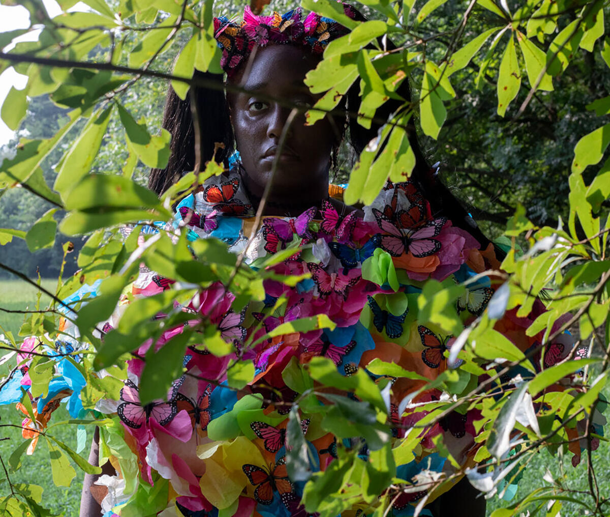 person looking at camera through leaves and wearing colorful butterflies all over their shirt. 