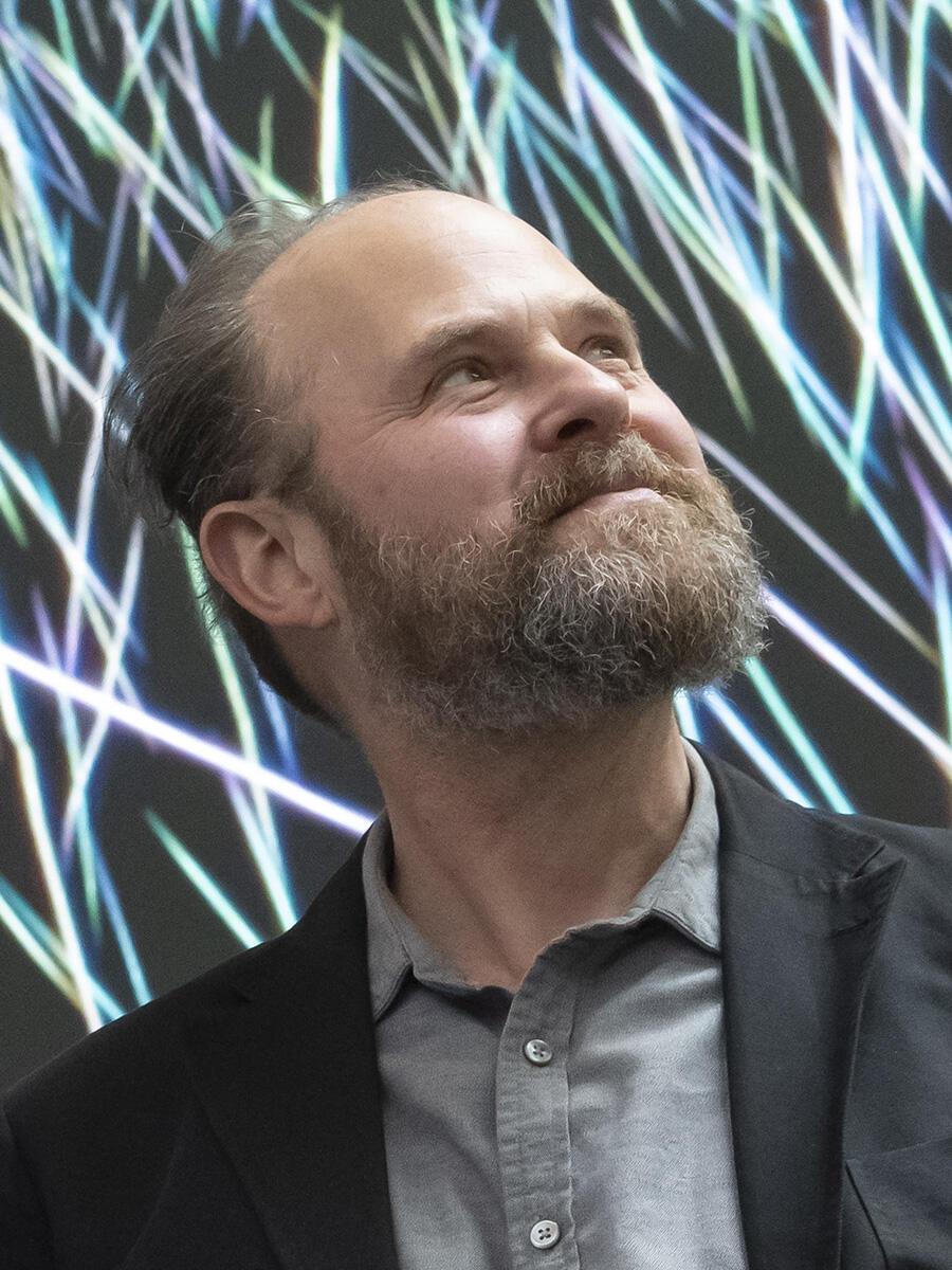 A bearded man looks to the side. A dark background is covered in white lines.