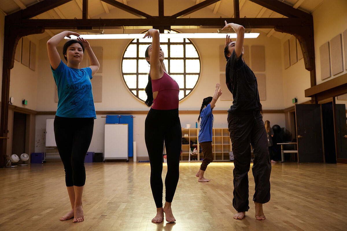 three dancers bend backwards in a room with a wood floor, beamed ceiling and large round window