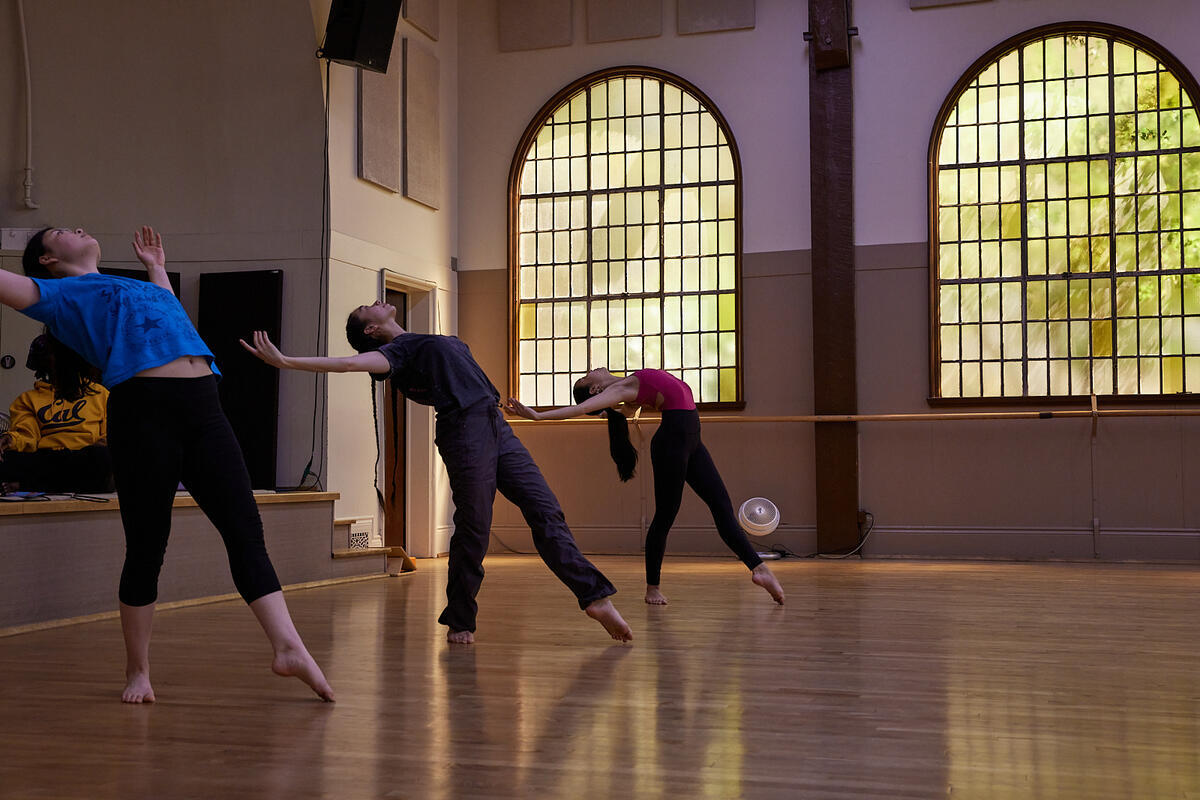 three dancers stand with outstretched arms in a room with a wood floor, beamed ceiling and large round window