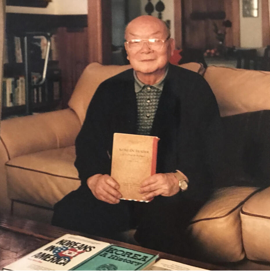 an older man sits on a couch holding a textbook. three other books are arrayed on the table in front of him