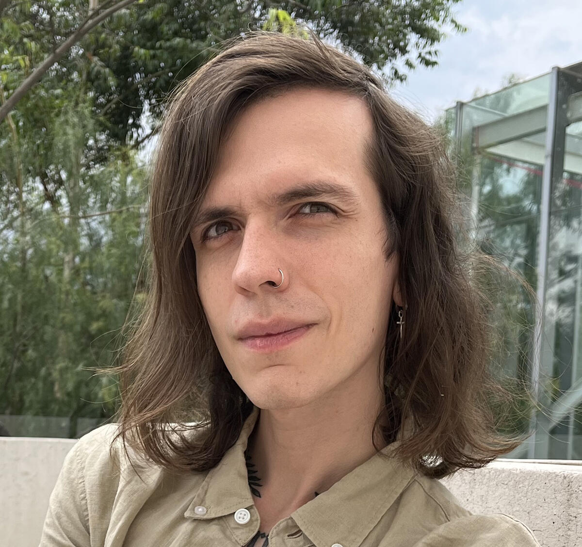 Close up of an individual with shoulder length hair and a nose ring outdoors in front of a glass building
