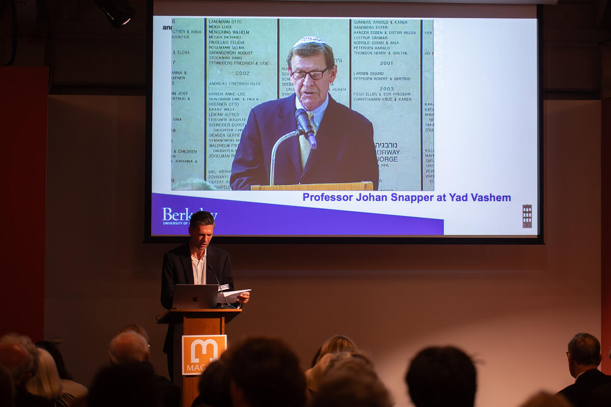 man speaking at podium; slide behind him shows image of man with text saying "Johann Snapper at Yad Vashem Memorial"