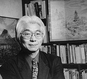 A man with white hair and glasses in front of a bookshelf and artworks.