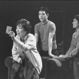 Three people in a black and white photo, one seated holding a card, another standing leaning forward, and the third standing to the side.