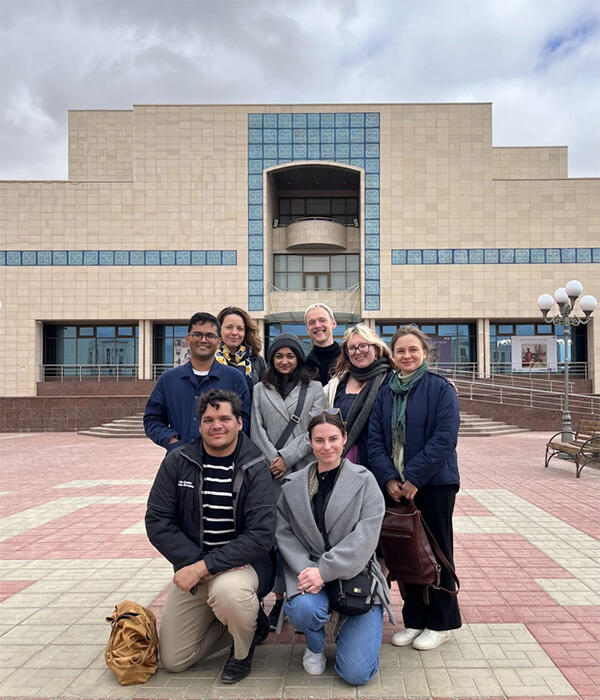 Eight people pose for a photo on a plaza in front of a tan and blue building