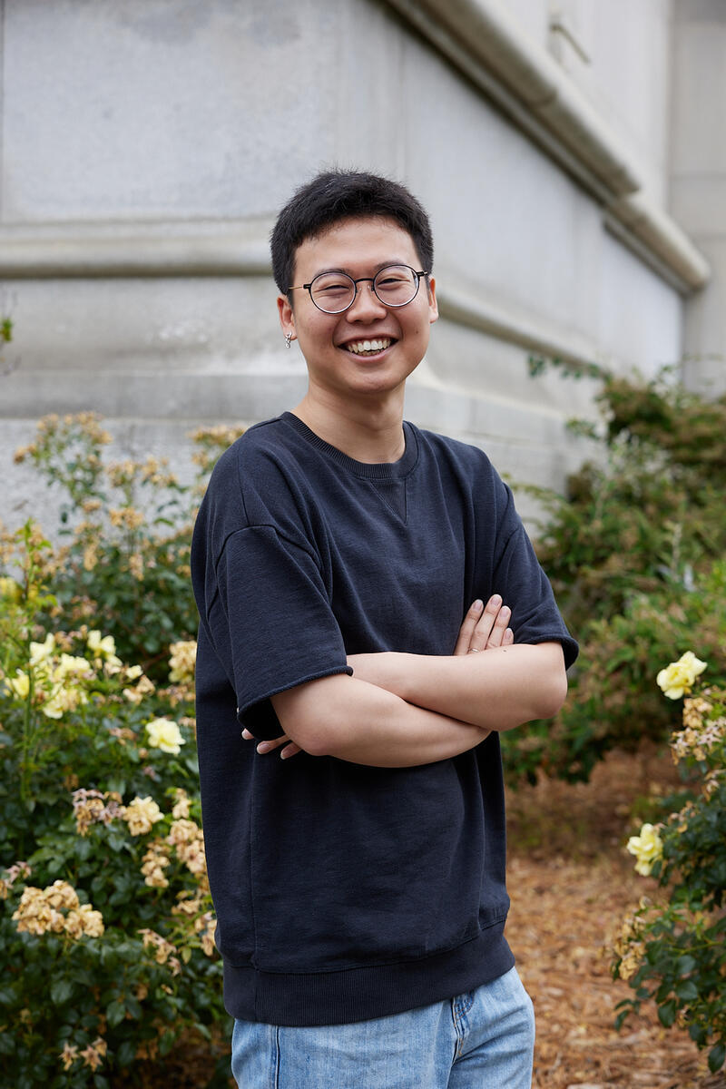 a smiling student with glasses and crossed arms stands in front of yellow rosebushes