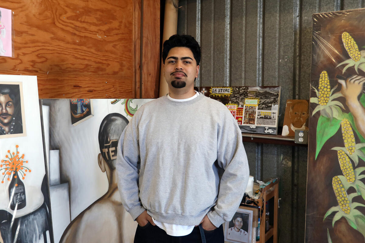 A man in a light gray sweater stands in his studio, with large paintings leaning against the wall in the background