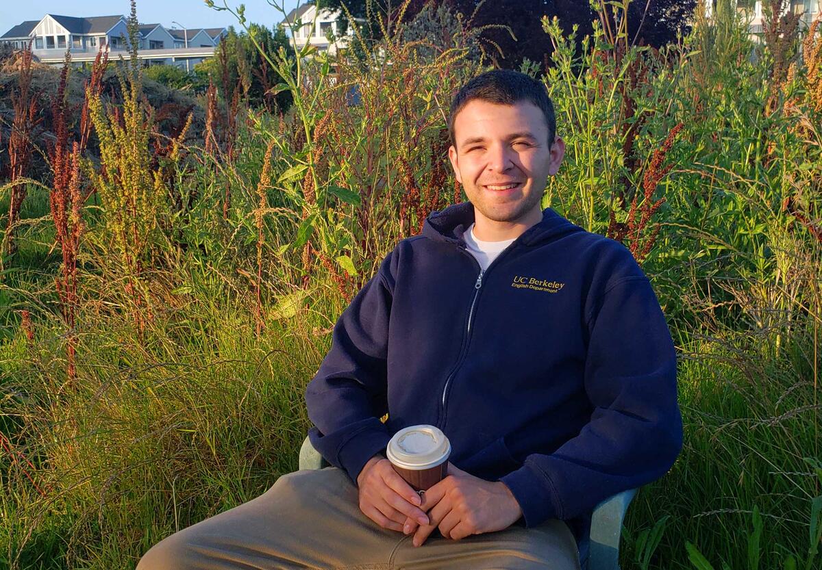 man wearing a UC Berkeley English sweatshirt sits in a chair in a garden