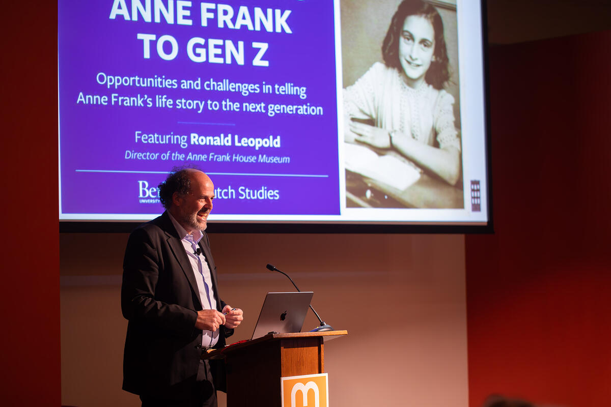 man speaking at podium with image of Anne Frank on screen behind him
