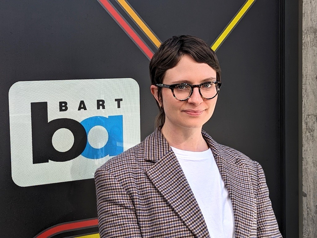 woman in professional attire with short hair and glasses stands in front of BART sign