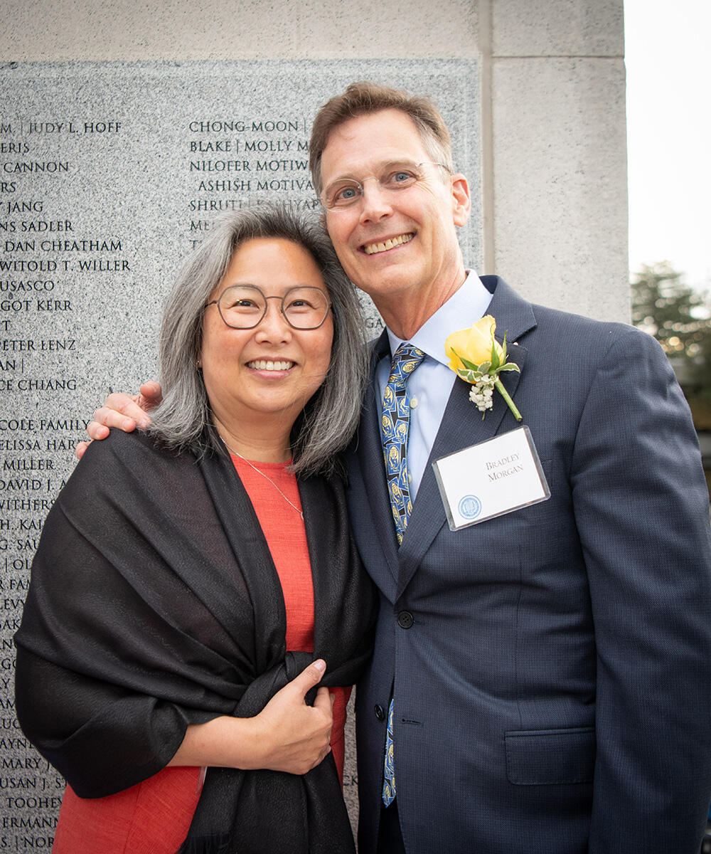 Brad Morgan and Julia Shin Morgan stand smiling in front of a granite wall engraved with names