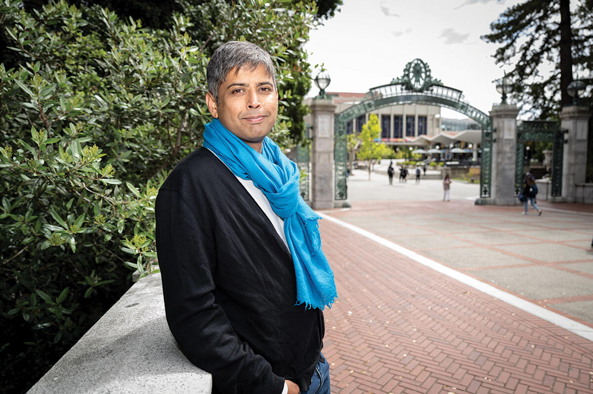 man with black sweater and blue scarf smiles at camera with Sather Gate in background